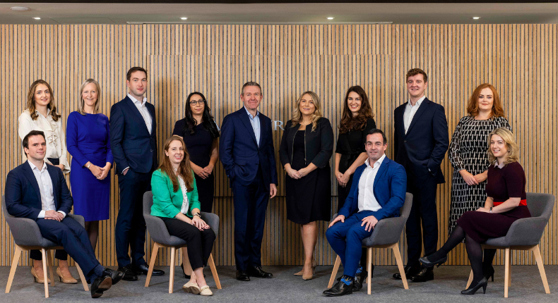 A group of people, some standing and some seated, in front of a slatted wooden wall.